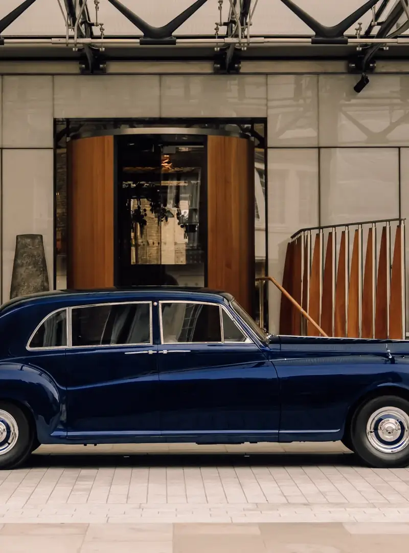 A classic dark blue luxury car is parked in front of a grand hotel entrance with a revolving wooden door and wide stone steps on either side.