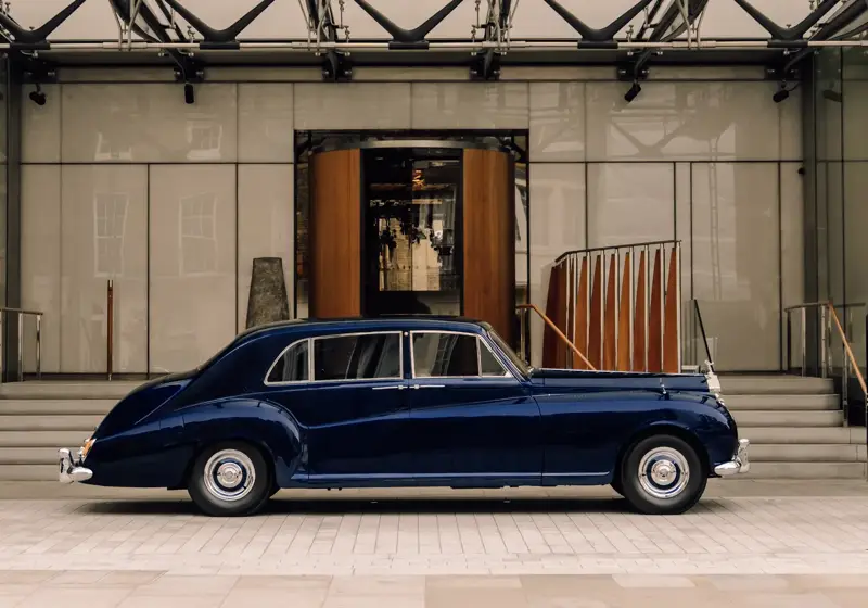A classic dark blue luxury car is parked in front of a grand hotel entrance with a revolving wooden door and wide stone steps on either side.