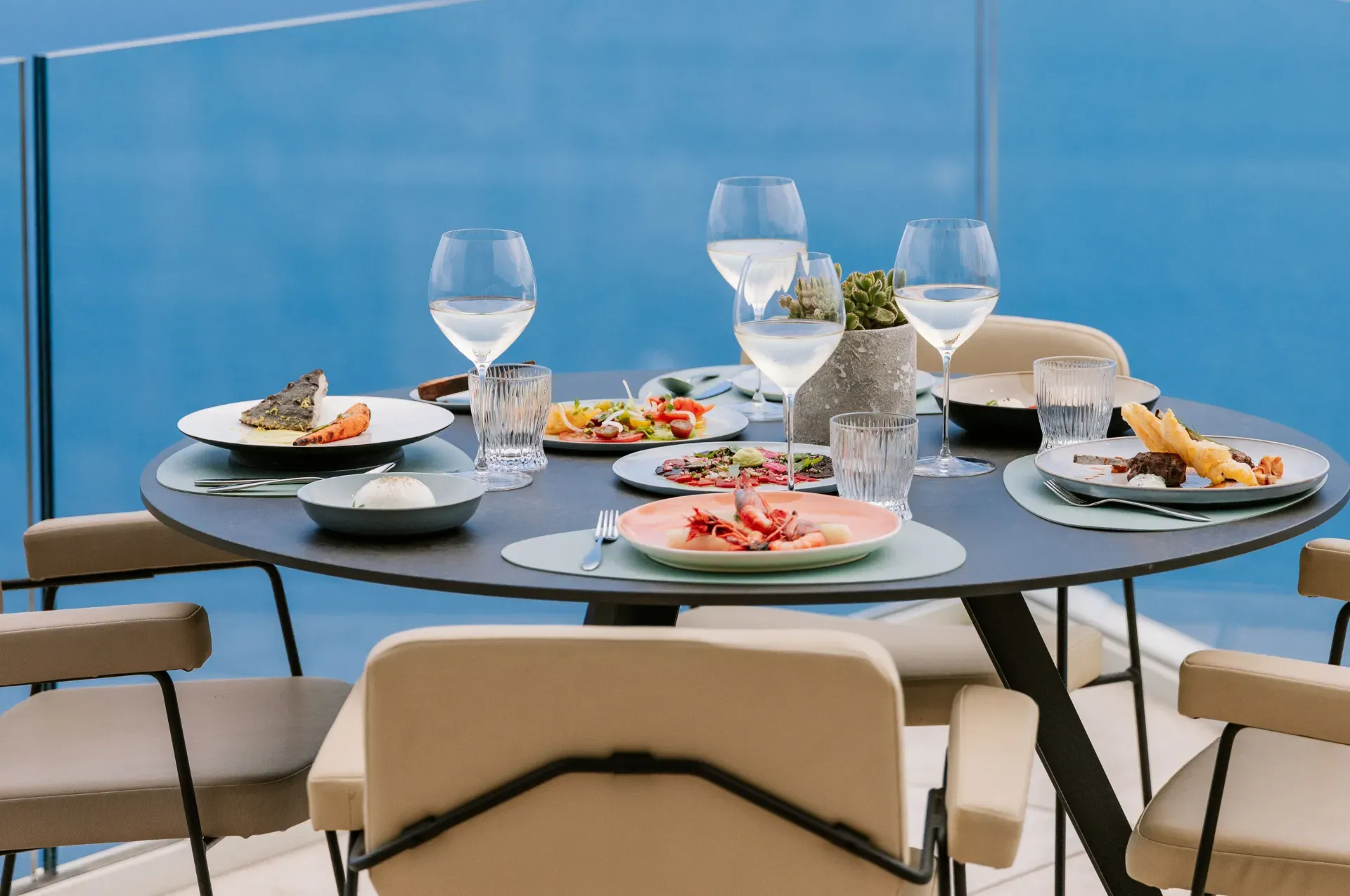 Table ronde dressée avec plats de fruits de mer et verres à vin près d’une baie vitrée avec vue sur la mer bleue.