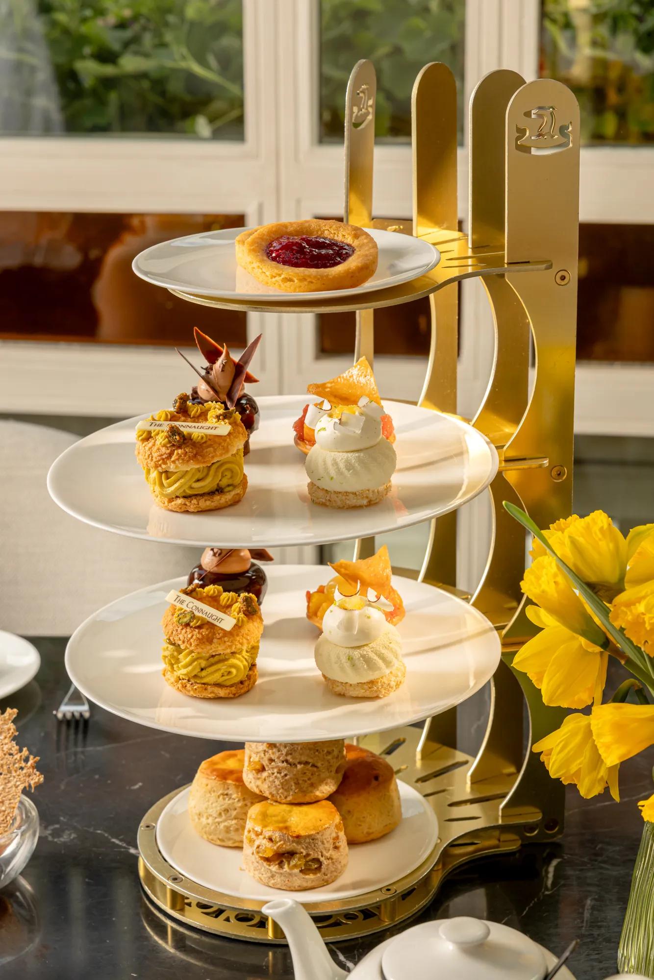 Four-tier afternoon tea stand with fruit tart, pastries, and scones beside yellow flowers.