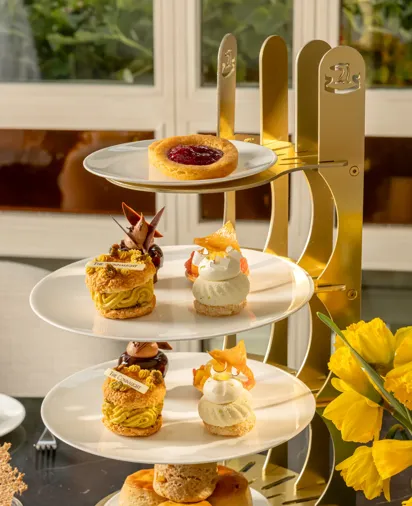 Four-tier afternoon tea stand with fruit tart, pastries, and scones beside yellow flowers.