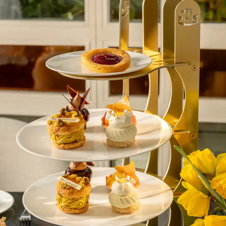 Four-tier afternoon tea stand with fruit tart, pastries, and scones beside yellow flowers.