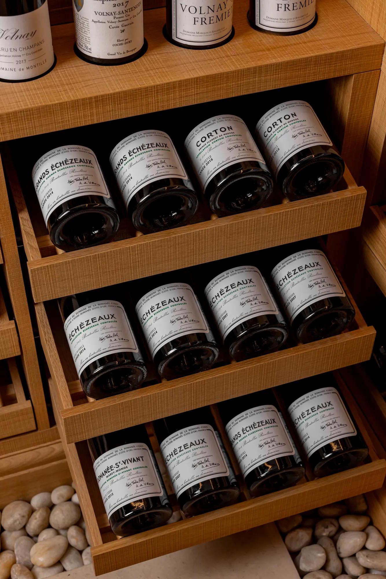 Wooden wine racks displaying horizontally stacked bottles of fine Burgundy wines, including Clos de Vougeot, Corton, and Romanée-Saint-Vivant, resting above smooth white pebbles on the floor.