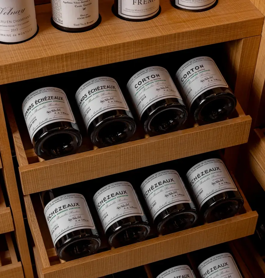 Wooden wine racks displaying horizontally stacked bottles of fine Burgundy wines, including Clos de Vougeot, Corton, and Romanée-Saint-Vivant, resting above smooth white pebbles on the floor.