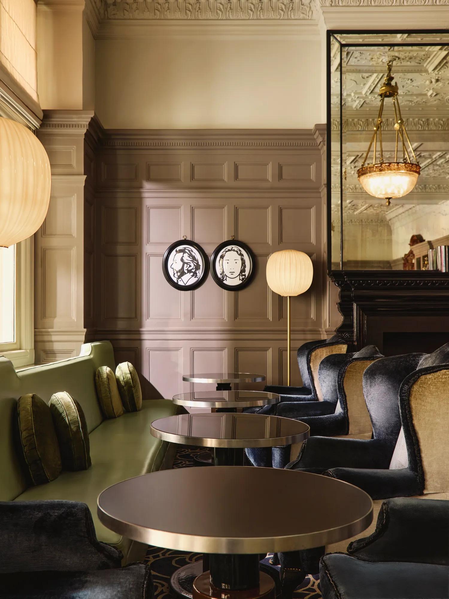 Elegant lounge seating with velvet chairs, round tables, and paneled walls decorated with framed portraits and warm lighting.