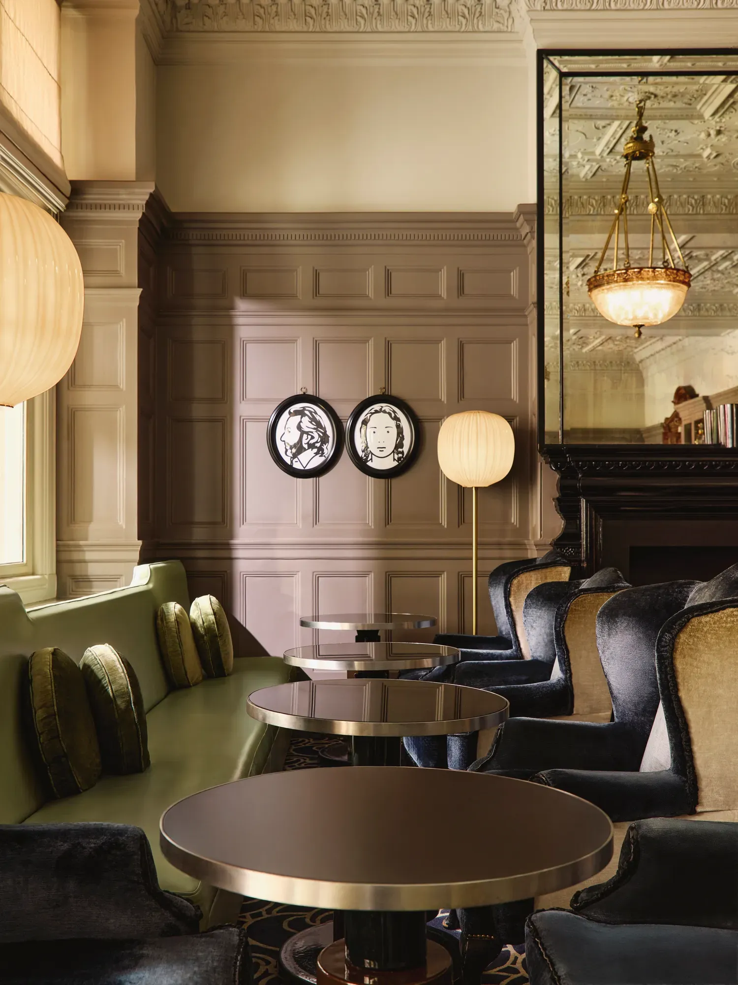 Elegant lounge seating with velvet chairs, round tables, and paneled walls decorated with framed portraits and warm lighting.