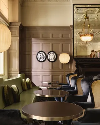 Elegant lounge seating with velvet chairs, round tables, and paneled walls decorated with framed portraits and warm lighting.