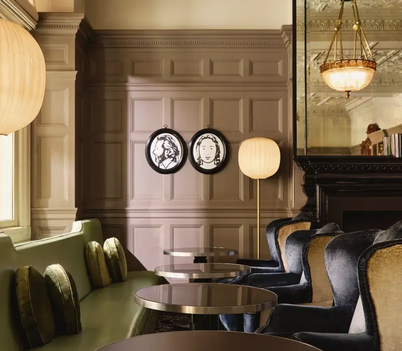 Elegant lounge seating with velvet chairs, round tables, and paneled walls decorated with framed portraits and warm lighting.