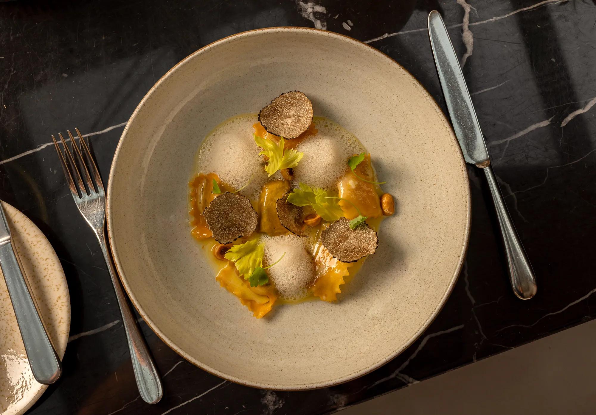 A dish of handmade ravioli topped with shaved truffle and delicate espuma, served in a wide ceramic bowl on a polished marble table.