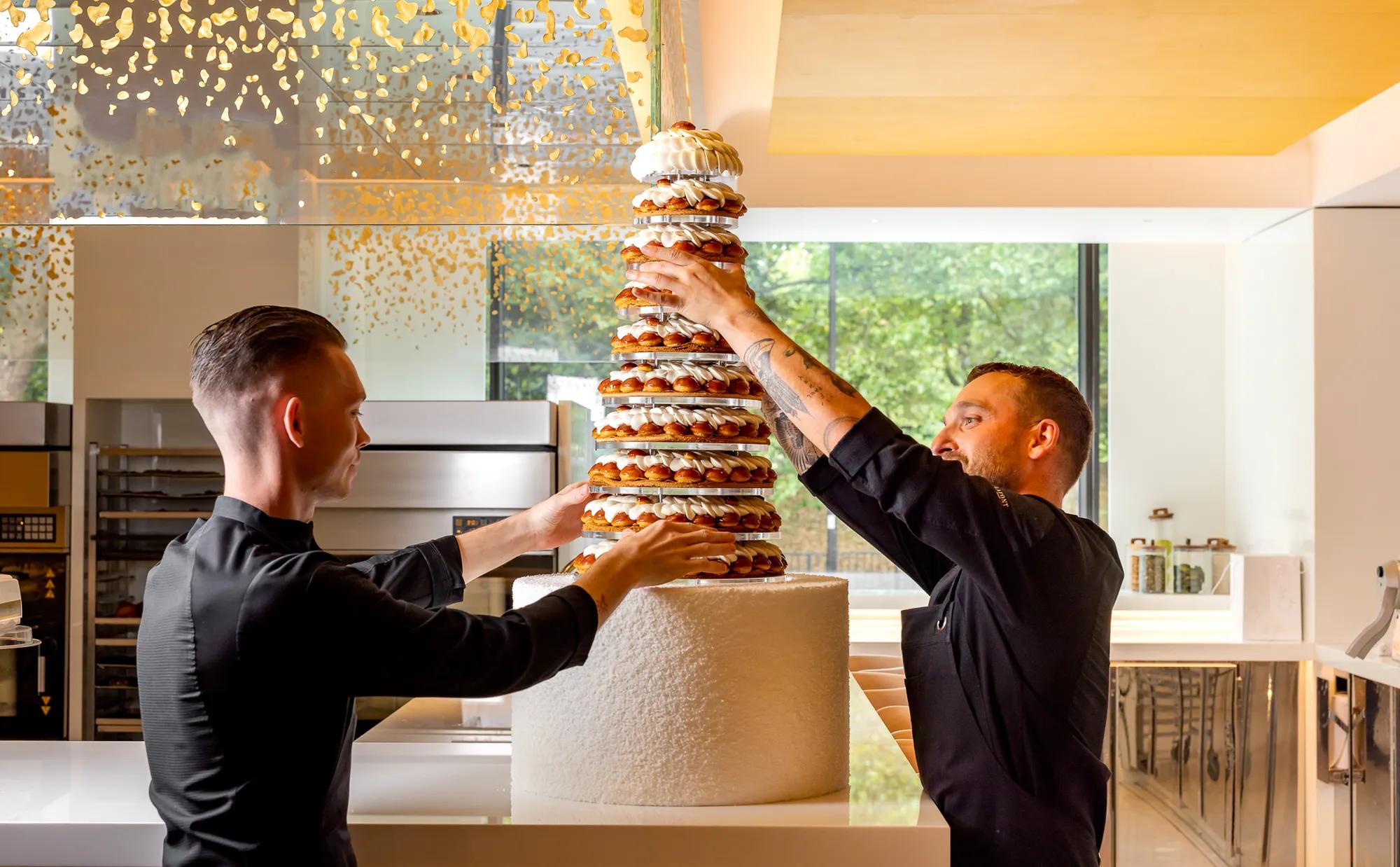 Pastry chefs assembling tall croquembouche wedding cake in modern kitchen with gold ceiling accents.