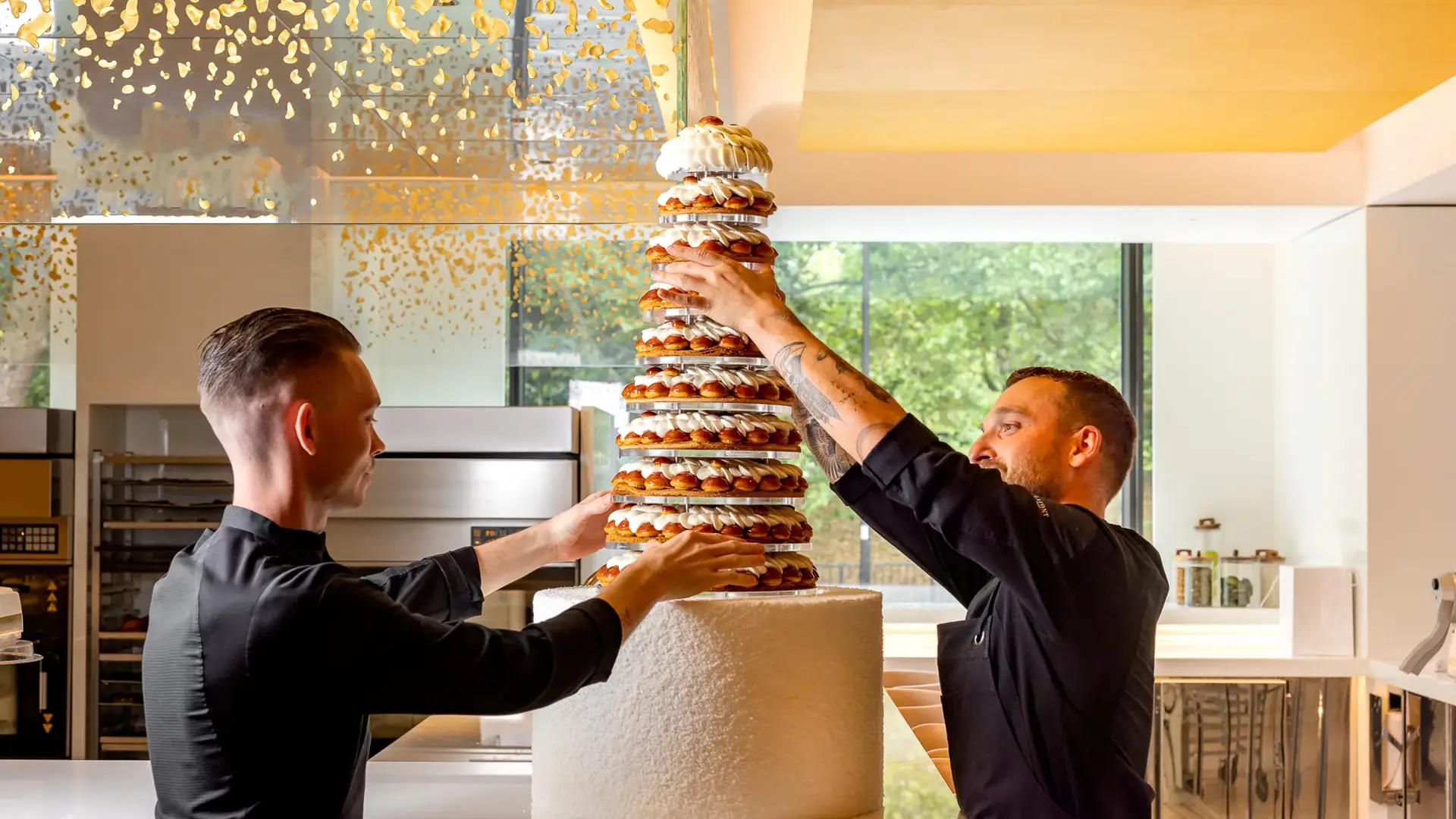 Pastry chefs assembling tall croquembouche wedding cake in modern kitchen with gold ceiling accents.