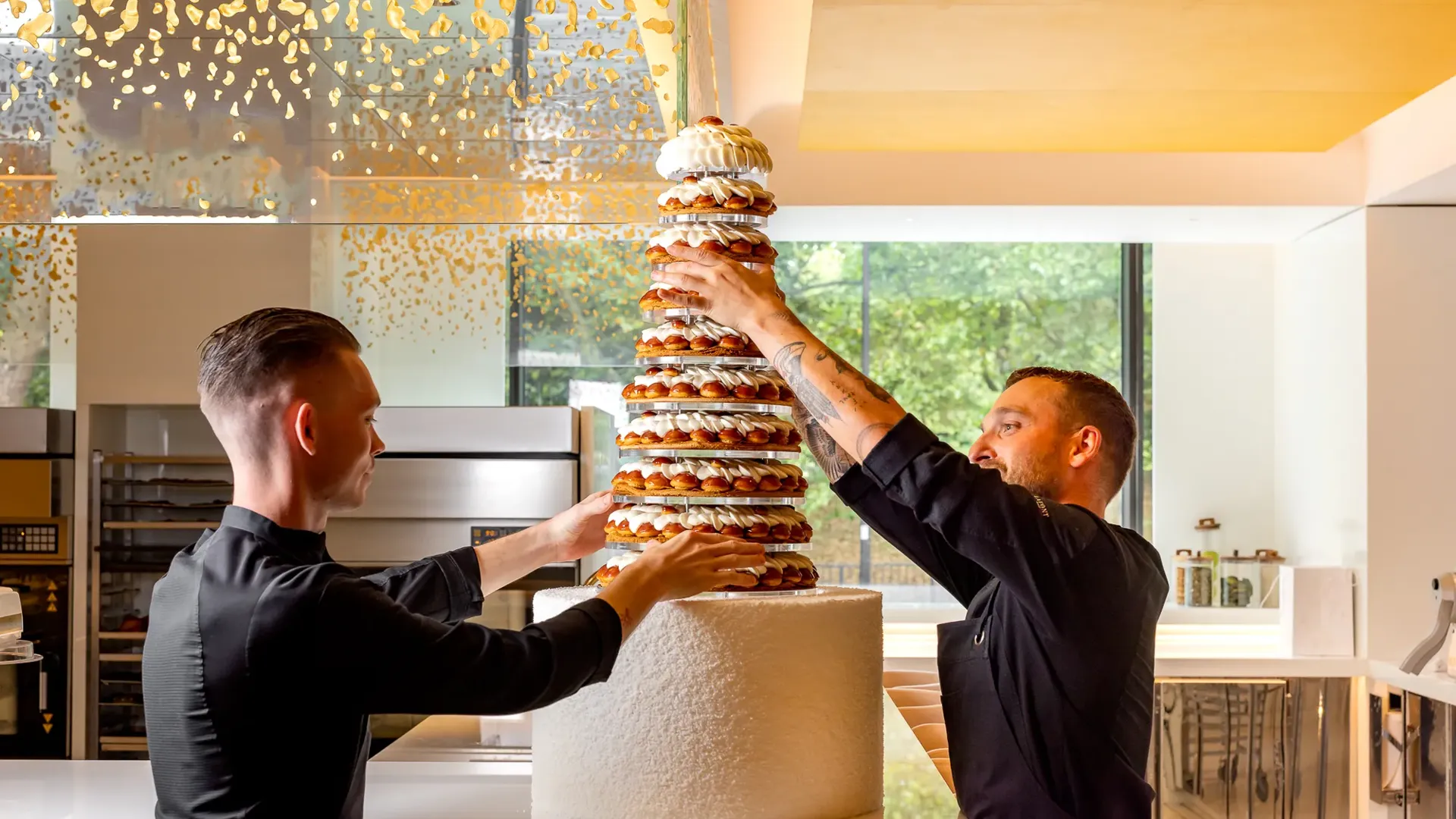 Pastry chefs assembling tall croquembouche wedding cake in modern kitchen with gold ceiling accents.
