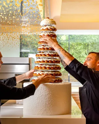 Pastry chefs assembling tall croquembouche wedding cake in modern kitchen with gold ceiling accents.