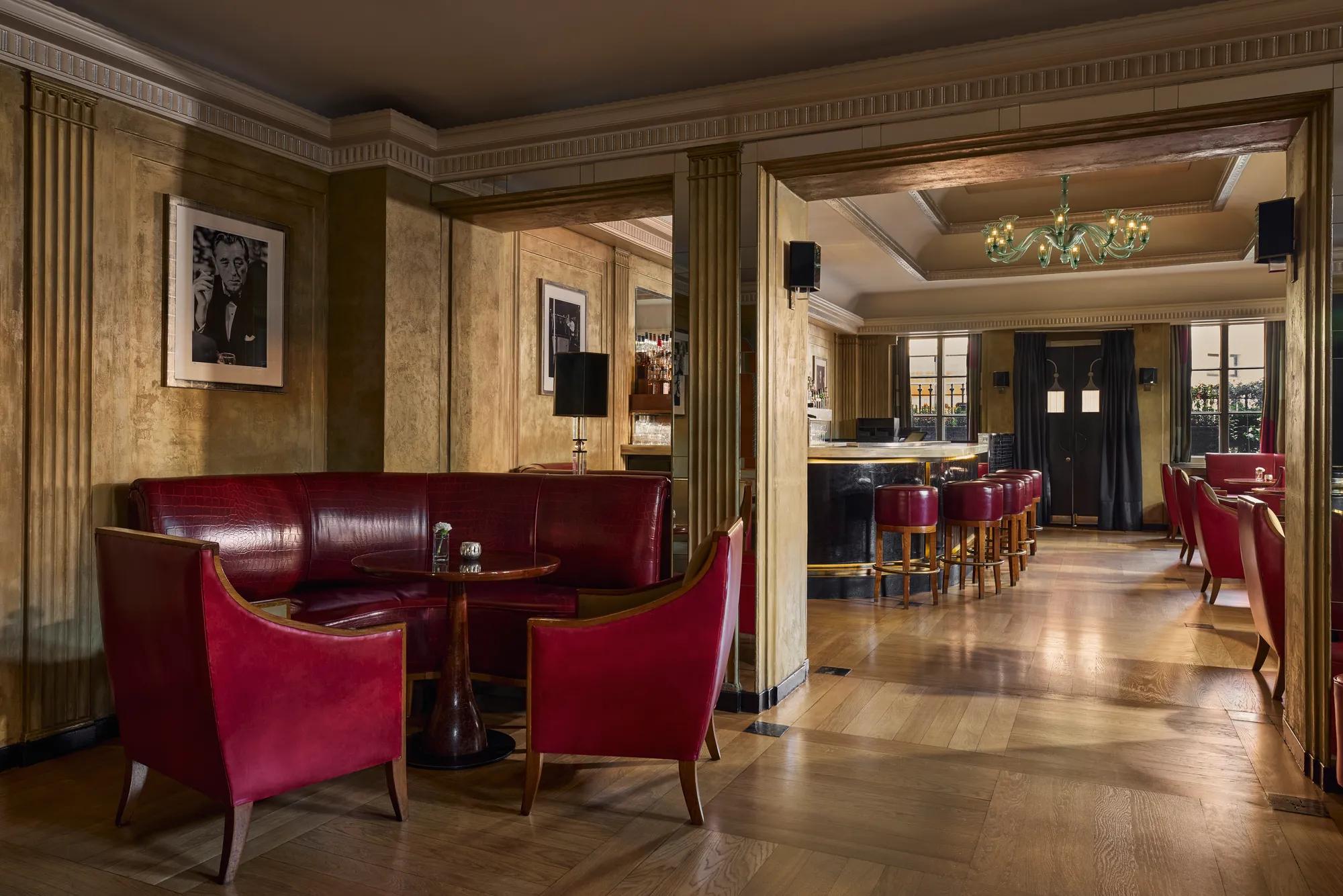 Elegant bar lounge featuring red leather seating, a round wooden table, and framed black-and-white portraits on golden-toned walls. The view extends to a polished bar area with high stools and a chandelier, illuminated by natural light from large windows.