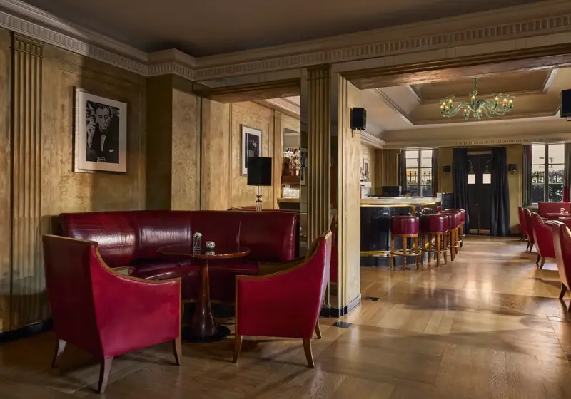 Elegant bar lounge featuring red leather seating, a round wooden table, and framed black-and-white portraits on golden-toned walls. The view extends to a polished bar area with high stools and a chandelier, illuminated by natural light from large windows.