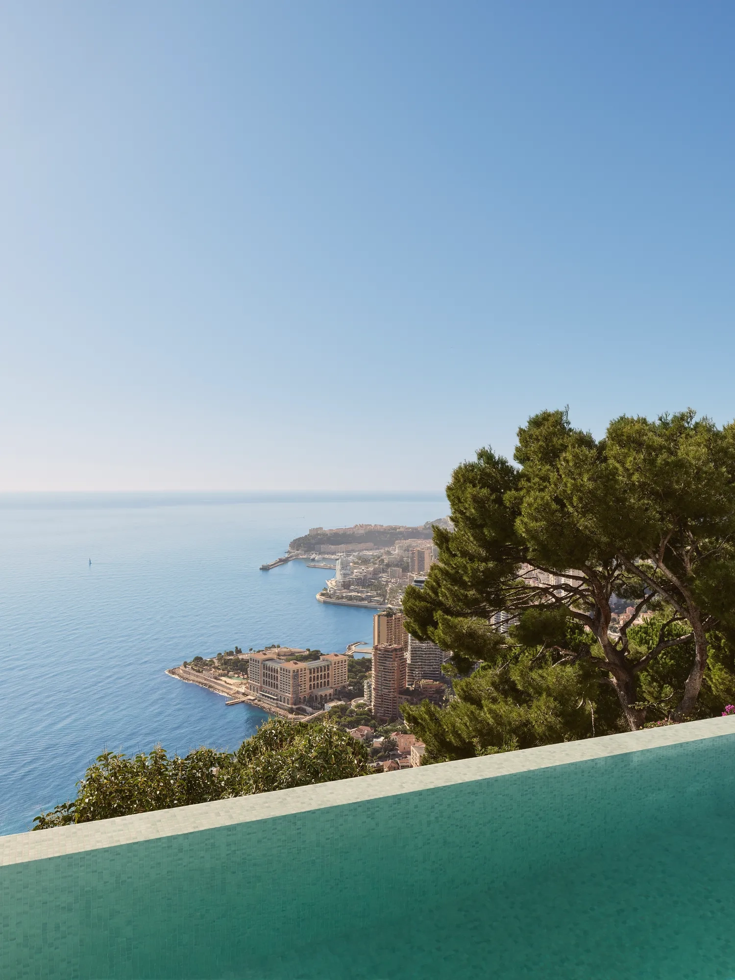 Infinity pool overlooking the sea and Monaco’s coastline, framed by lush trees under a clear blue sky.