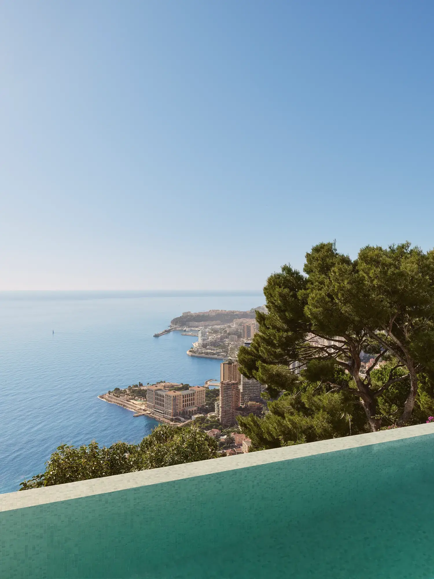 Infinity pool overlooking the sea and Monaco’s coastline, framed by lush trees under a clear blue sky.