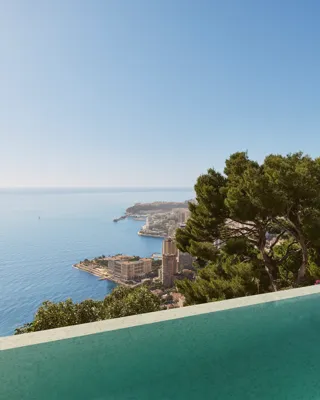 Infinity pool overlooking the sea and Monaco’s coastline, framed by lush trees under a clear blue sky.