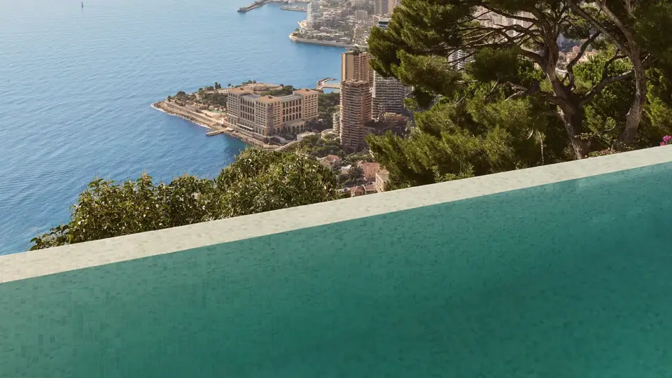 Piscine à débordement surplombant la mer et le littoral monégasque, encadrée par des arbres luxuriants sous un ciel bleu clair.