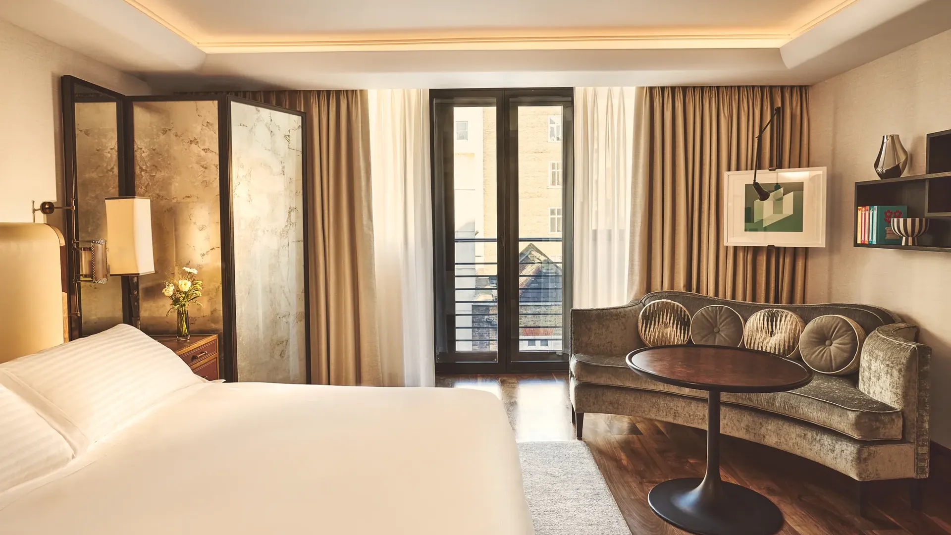 Warm, modern bedroom with a large bed, curved sofa and round table, and floor-to-ceiling windows with sheer and blackout curtains.