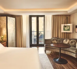 Warm, modern bedroom with a large bed, curved sofa and round table, and floor-to-ceiling windows with sheer and blackout curtains.