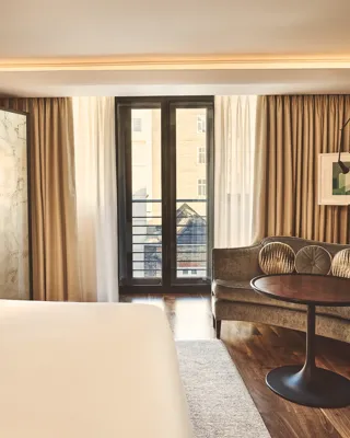 Warm, modern bedroom with a large bed, curved sofa and round table, and floor-to-ceiling windows with sheer and blackout curtains.