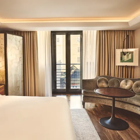 Warm, modern bedroom with a large bed, curved sofa and round table, and floor-to-ceiling windows with sheer and blackout curtains.