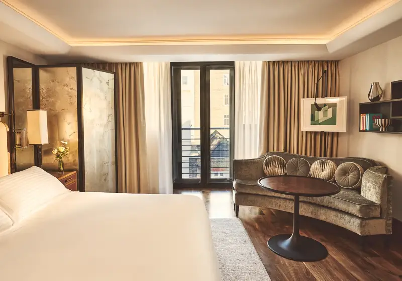 Warm, modern bedroom with a large bed, curved sofa and round table, and floor-to-ceiling windows with sheer and blackout curtains.