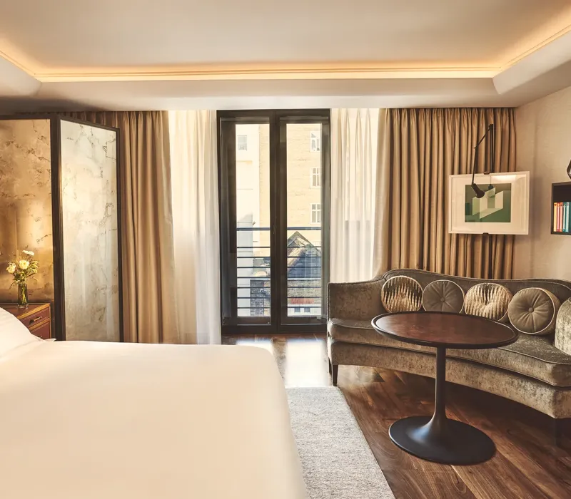 Warm, modern bedroom with a large bed, curved sofa and round table, and floor-to-ceiling windows with sheer and blackout curtains.