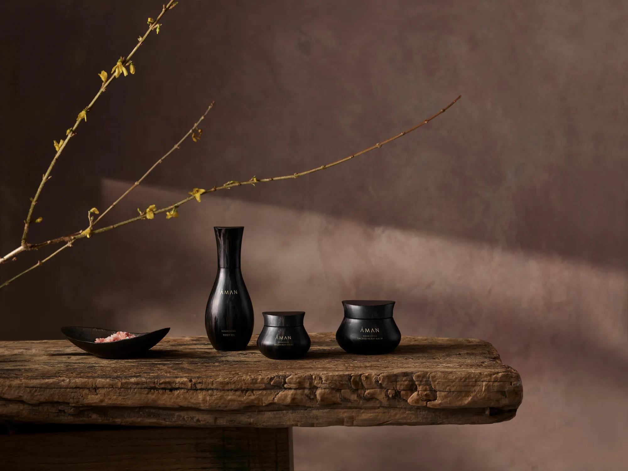Three black Aman skincare jars displayed on a rustic wooden table beneath a long budding branch.