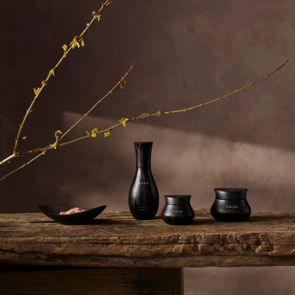 Three black Aman skincare jars displayed on a rustic wooden table beneath a long budding branch.