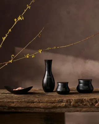 Three black Aman skincare jars displayed on a rustic wooden table beneath a long budding branch.
