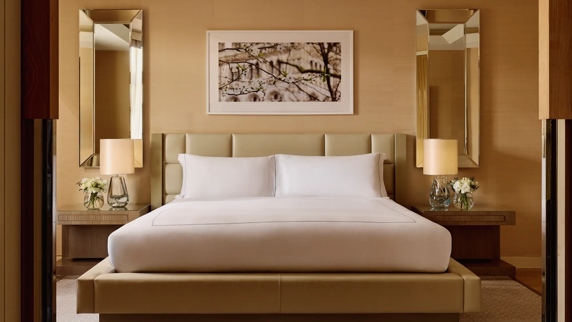 Elegant bedroom with a king bed, cream padded headboard, symmetrical mirrored wall panels, bedside tables with lamps and flowers, and a framed artwork above the bed.