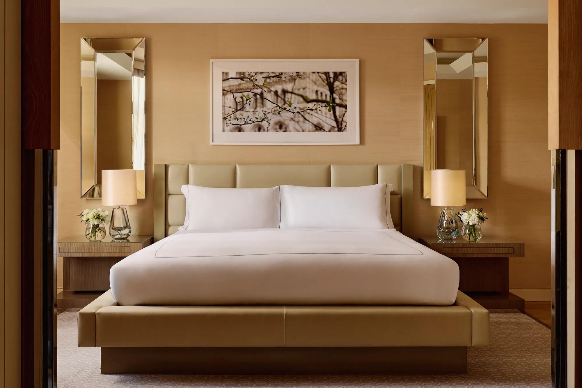 Elegant bedroom with a king bed, cream padded headboard, symmetrical mirrored wall panels, bedside tables with lamps and flowers, and a framed artwork above the bed.