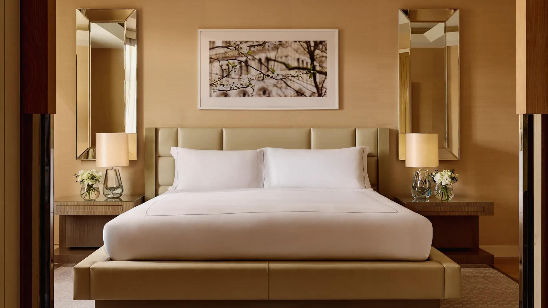 Elegant bedroom with a king bed, cream padded headboard, symmetrical mirrored wall panels, bedside tables with lamps and flowers, and a framed artwork above the bed.