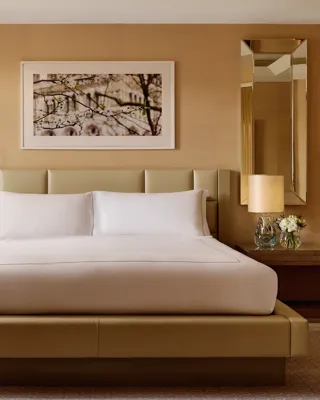 Elegant bedroom with a king bed, cream padded headboard, symmetrical mirrored wall panels, bedside tables with lamps and flowers, and a framed artwork above the bed.