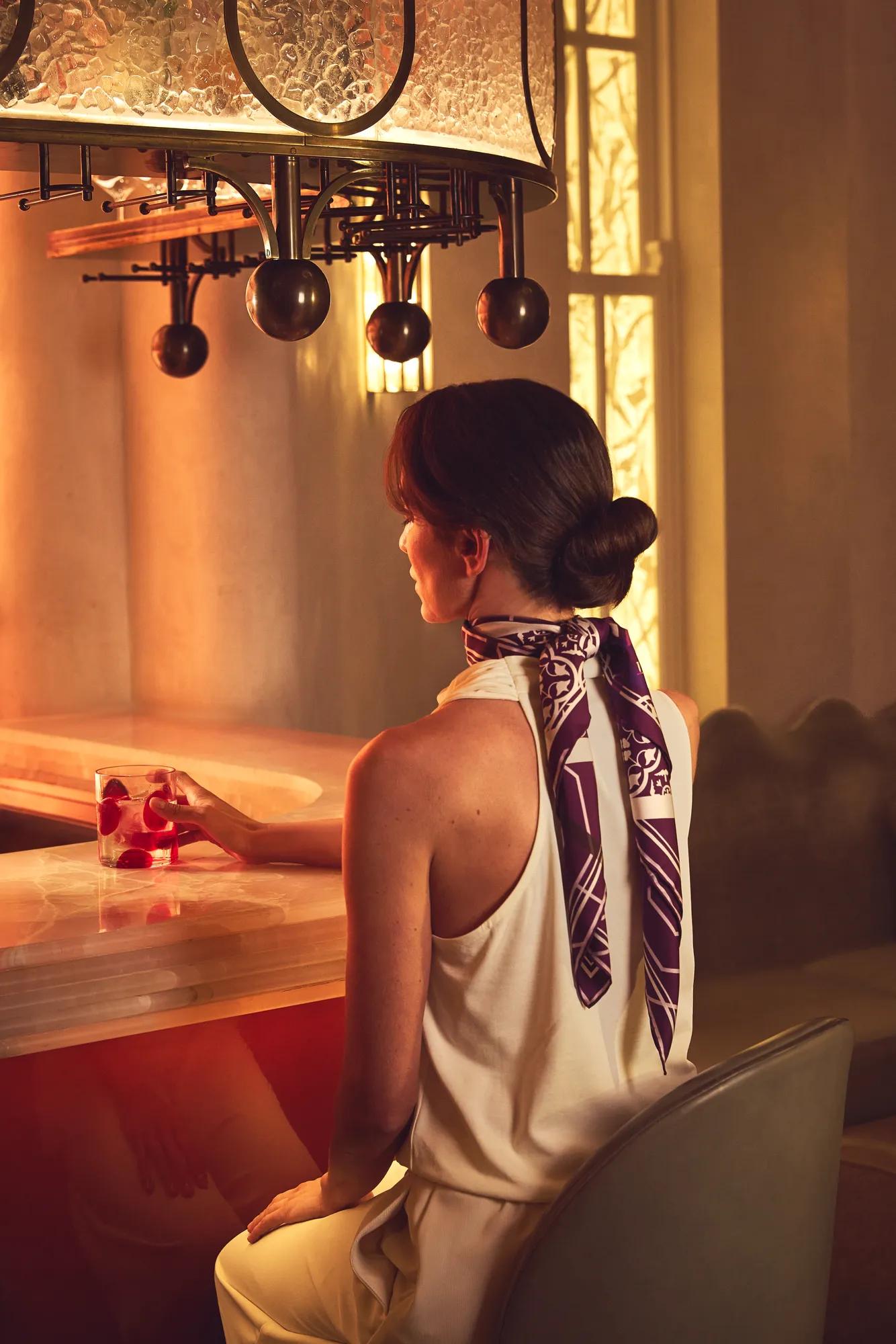 Woman seated at softly lit bar, holding red cocktail, wearing sleeveless top and silk scarf, with warm ambient lighting and decorative overhead fixture.
