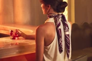Woman seated at softly lit bar, holding red cocktail, wearing sleeveless top and silk scarf, with warm ambient lighting and decorative overhead fixture.