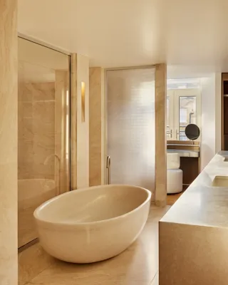 A warm-toned stone bathroom with an oval freestanding tub, glass shower, and floor-to-ceiling windows creating a serene, light-filled space.