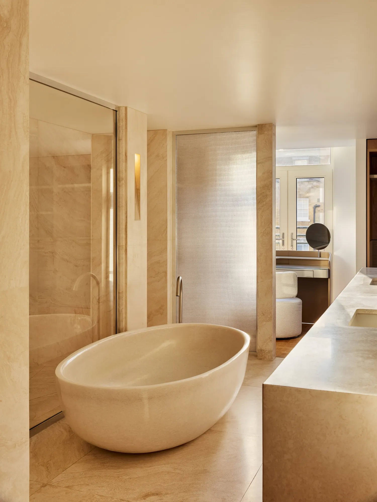 A warm-toned stone bathroom with an oval freestanding tub, glass shower, and floor-to-ceiling windows creating a serene, light-filled space.