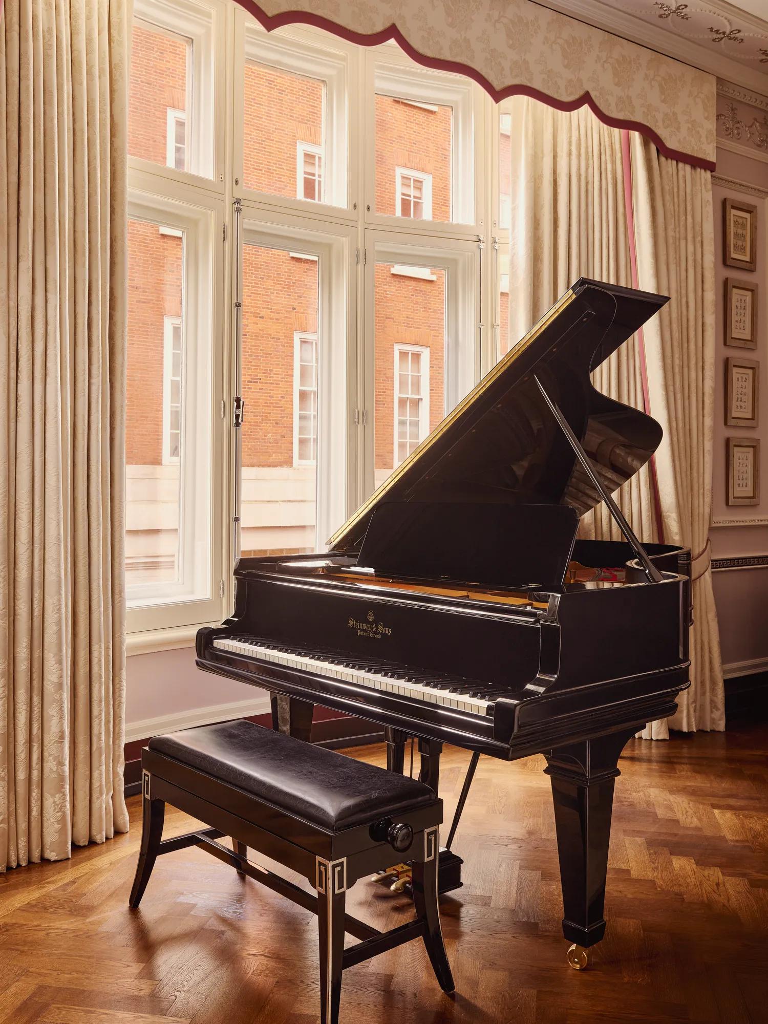 Black grand piano and bench positioned by tall curtained windows overlooking a red-brick building.