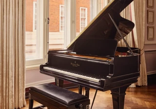 Black grand piano and bench positioned by tall curtained windows overlooking a red-brick building.