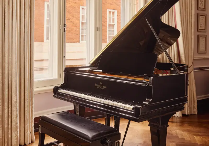 Black grand piano and bench positioned by tall curtained windows overlooking a red-brick building.