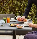 Server placing croissants on table with fruit plate, juices, tea set, jams, and pastries.