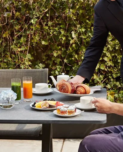 Server placing croissants on table with fruit plate, juices, tea set, jams, and pastries.