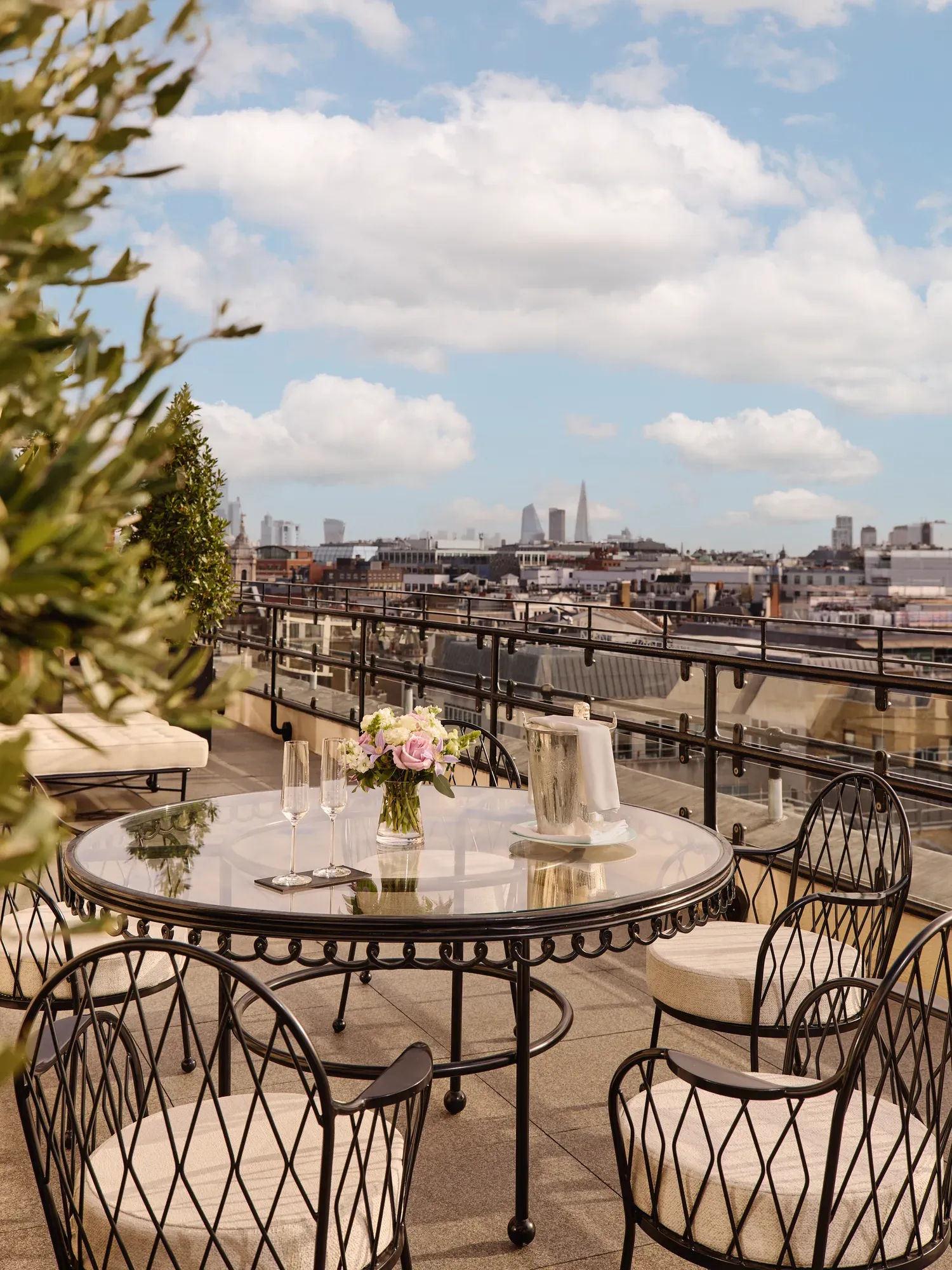 Rooftop terrace with a round glass dining table and black wrought-iron chairs, set with champagne glasses and flowers, overlooking London’s skyline under a partly cloudy sky.