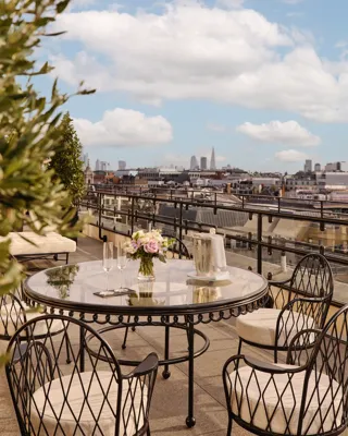Rooftop terrace with a round glass dining table and black wrought-iron chairs, set with champagne glasses and flowers, overlooking London’s skyline under a partly cloudy sky.