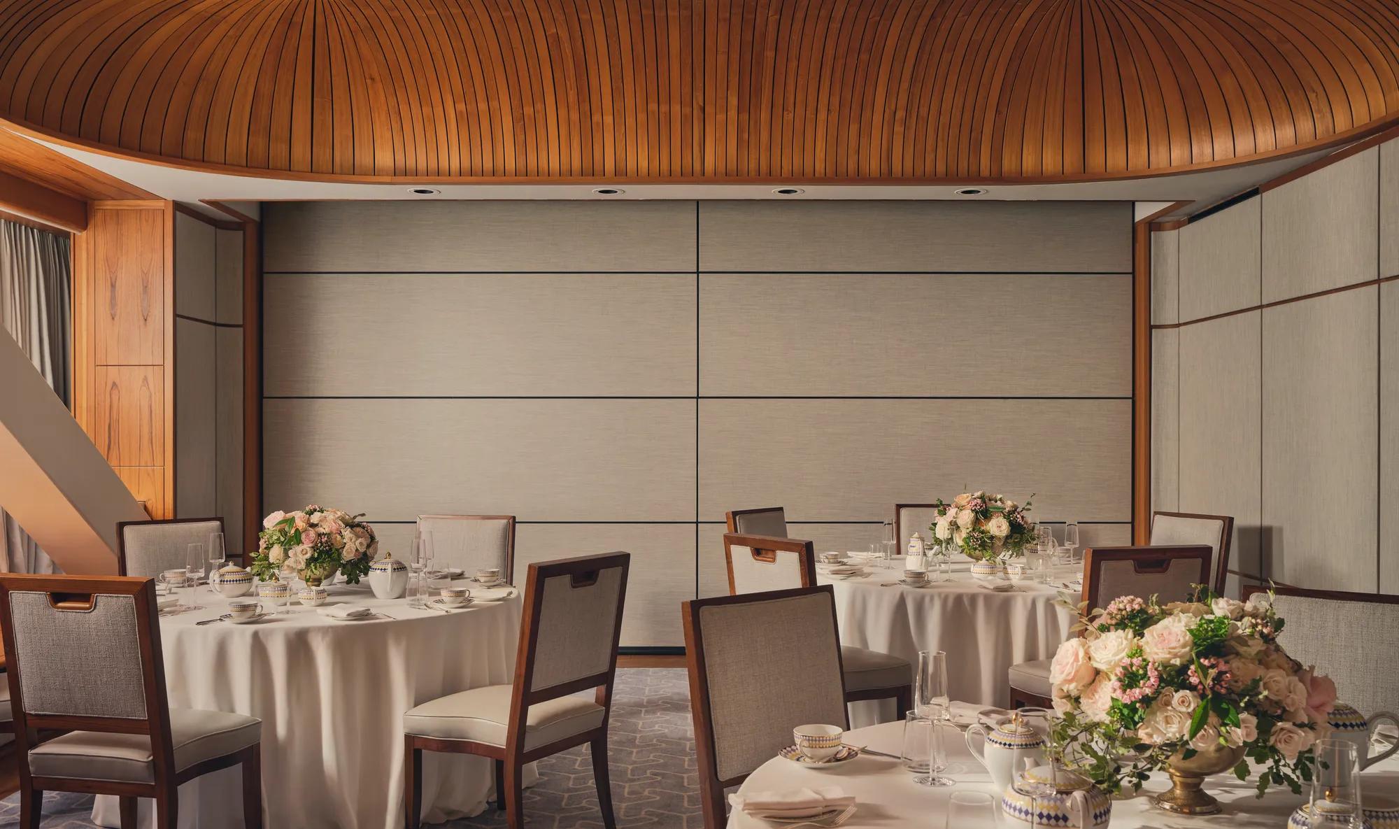 Intimate event room with round tables, floral centerpieces, fine china, and wood-panel ceiling under soft natural light.