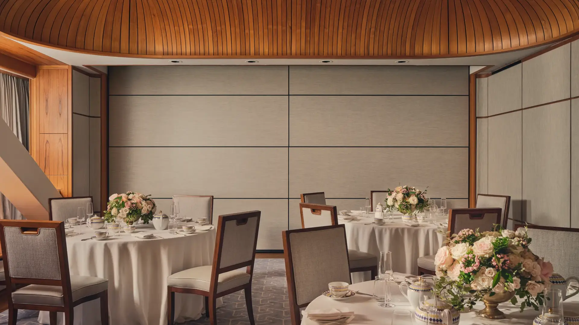 Intimate event room with round tables, floral centerpieces, fine china, and wood-panel ceiling under soft natural light.