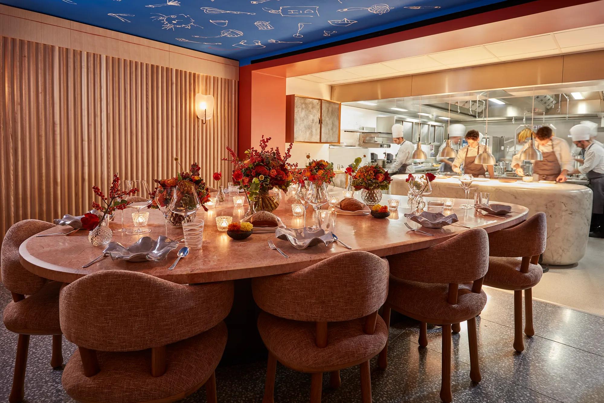 Candlelit chef’s table set with flowers beside an open kitchen where chefs prepare dishes.